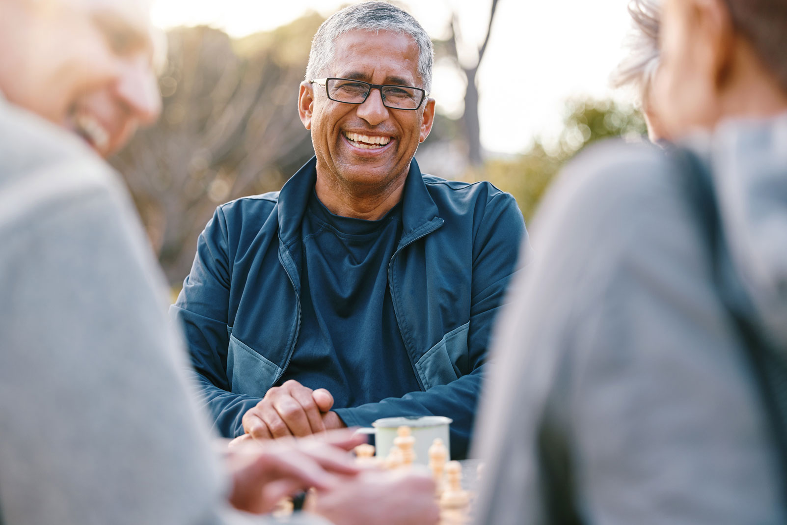 Man with glasses smiling while playing chess with friends outdoors.