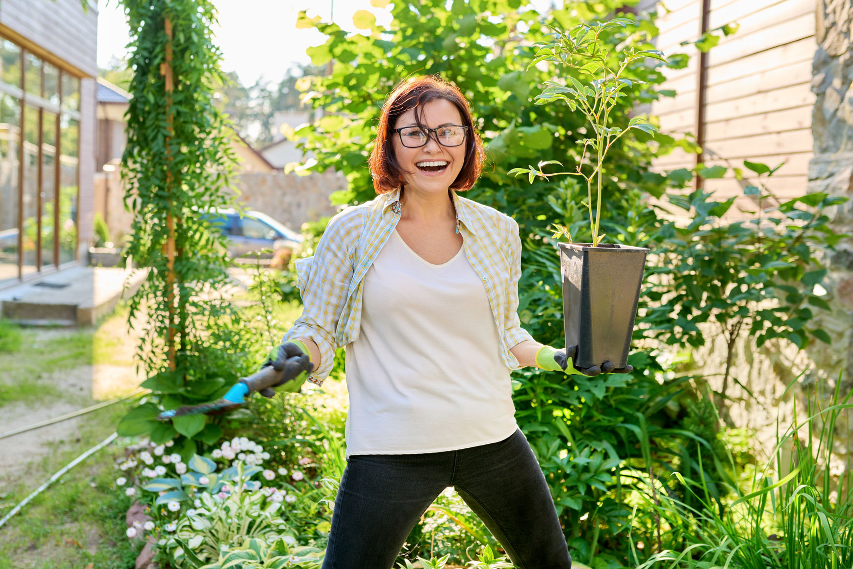 Woman in casual clothes smiling while holding a plant in a garden.