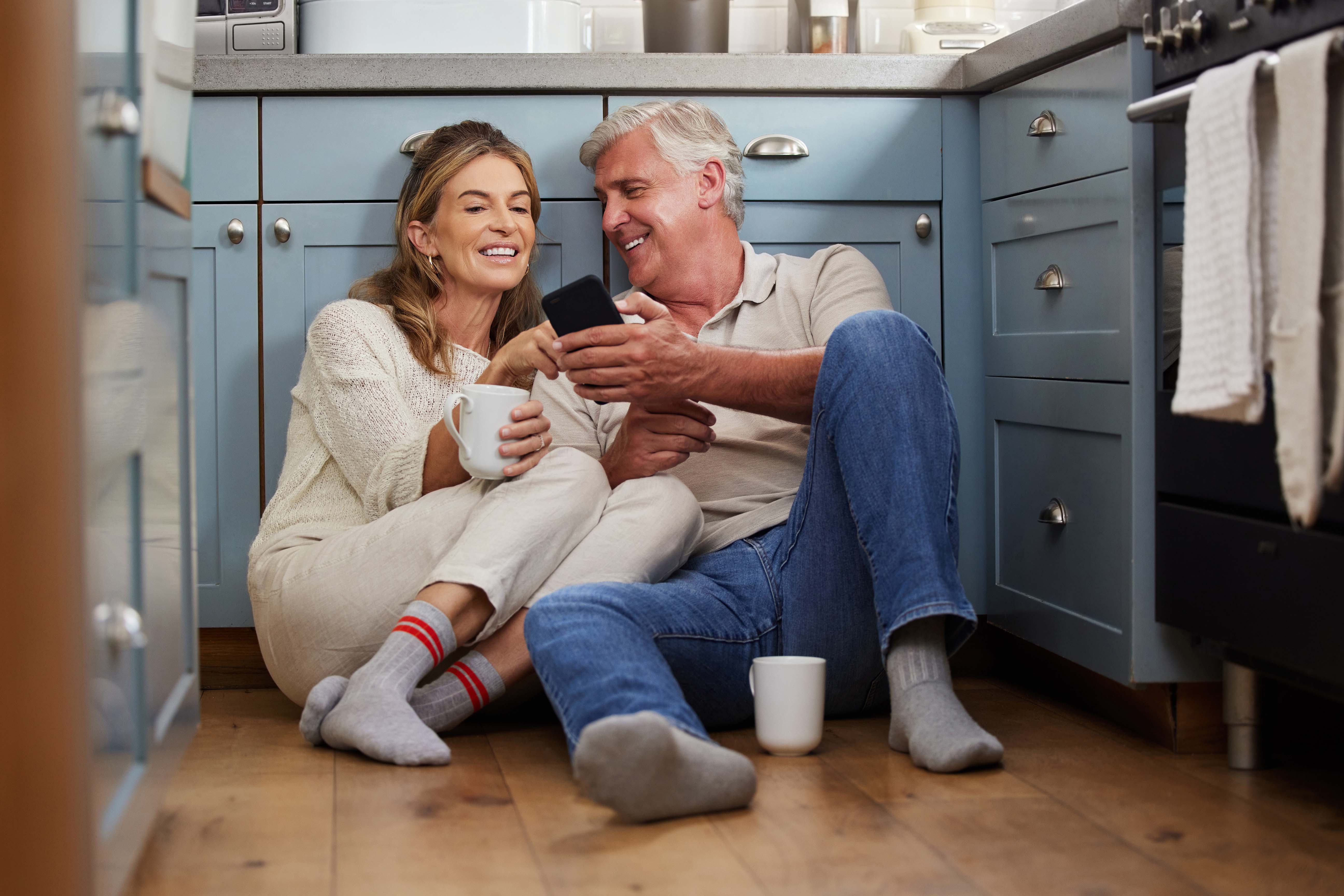 A couple sitting in the kitchen on the floor casually smiling and reading on the phone