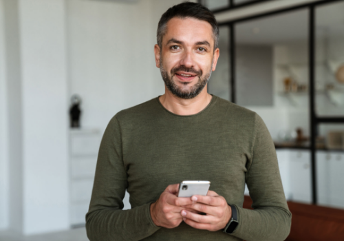 Man in a green sweater holding a smartphone and smiling indoors.