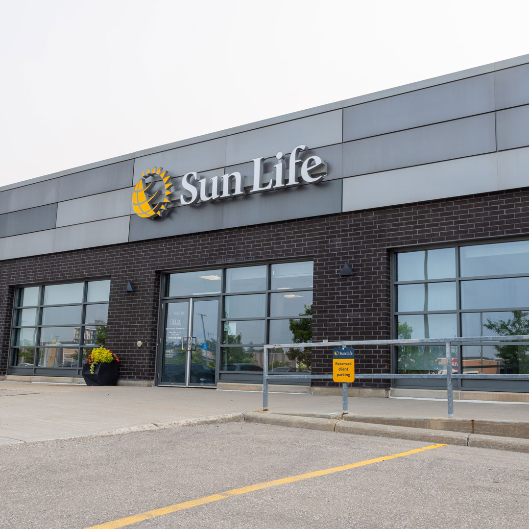 Wide view of the Sun Life office building from the parking lot, with the Sun Life logo above a dark brick storefront. The glass entrance door shows the Izzio Financial name, and yellow reserved client parking signs stand to the right.
