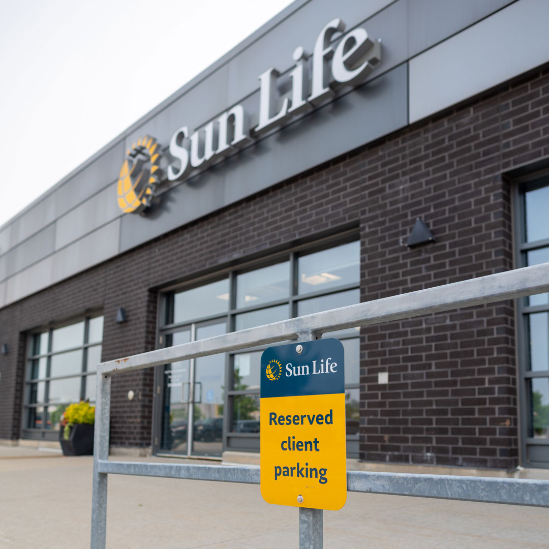 Close-up of a yellow and navy 'Reserved client parking' sign on a metal railing, with the Sun Life logo and brick storefront blurred behind it.