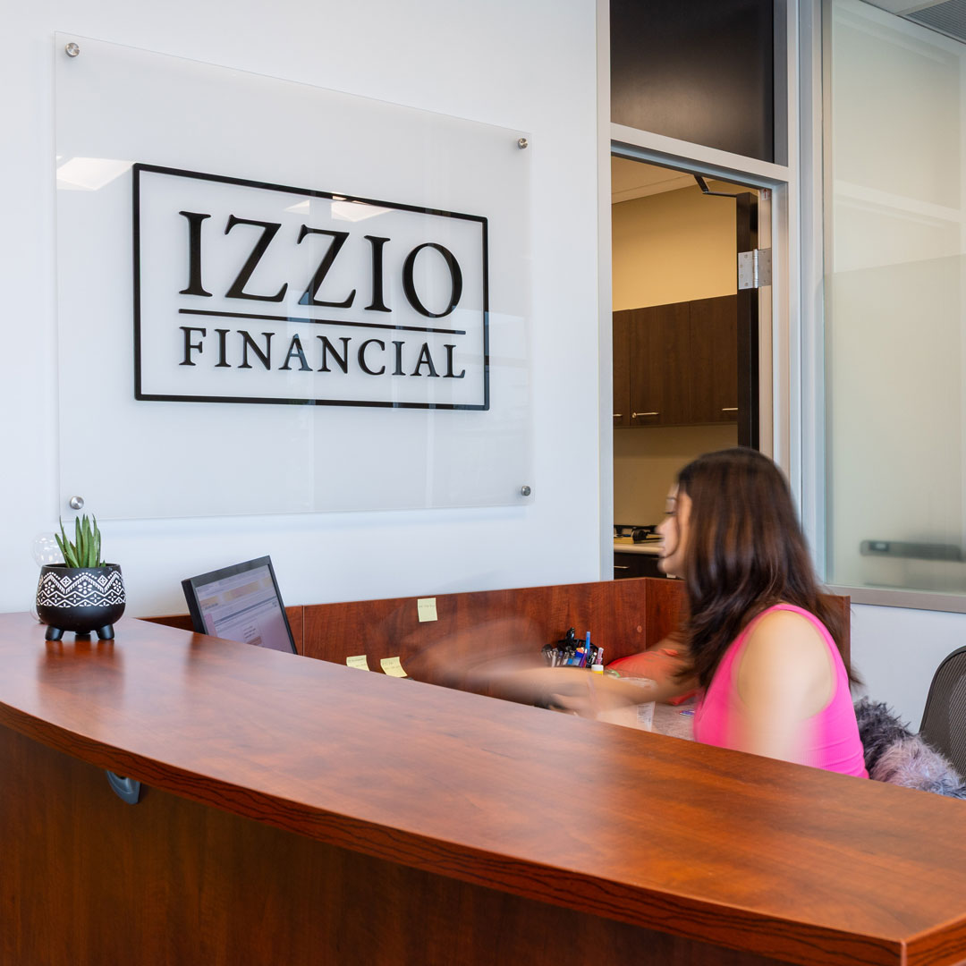 Reception area with the Izzio Financial logo on a frosted glass panel above a curved wood desk. A receptionist in a pink top sits at the desk, and a small plant in a patterned black pot sits on the counter.