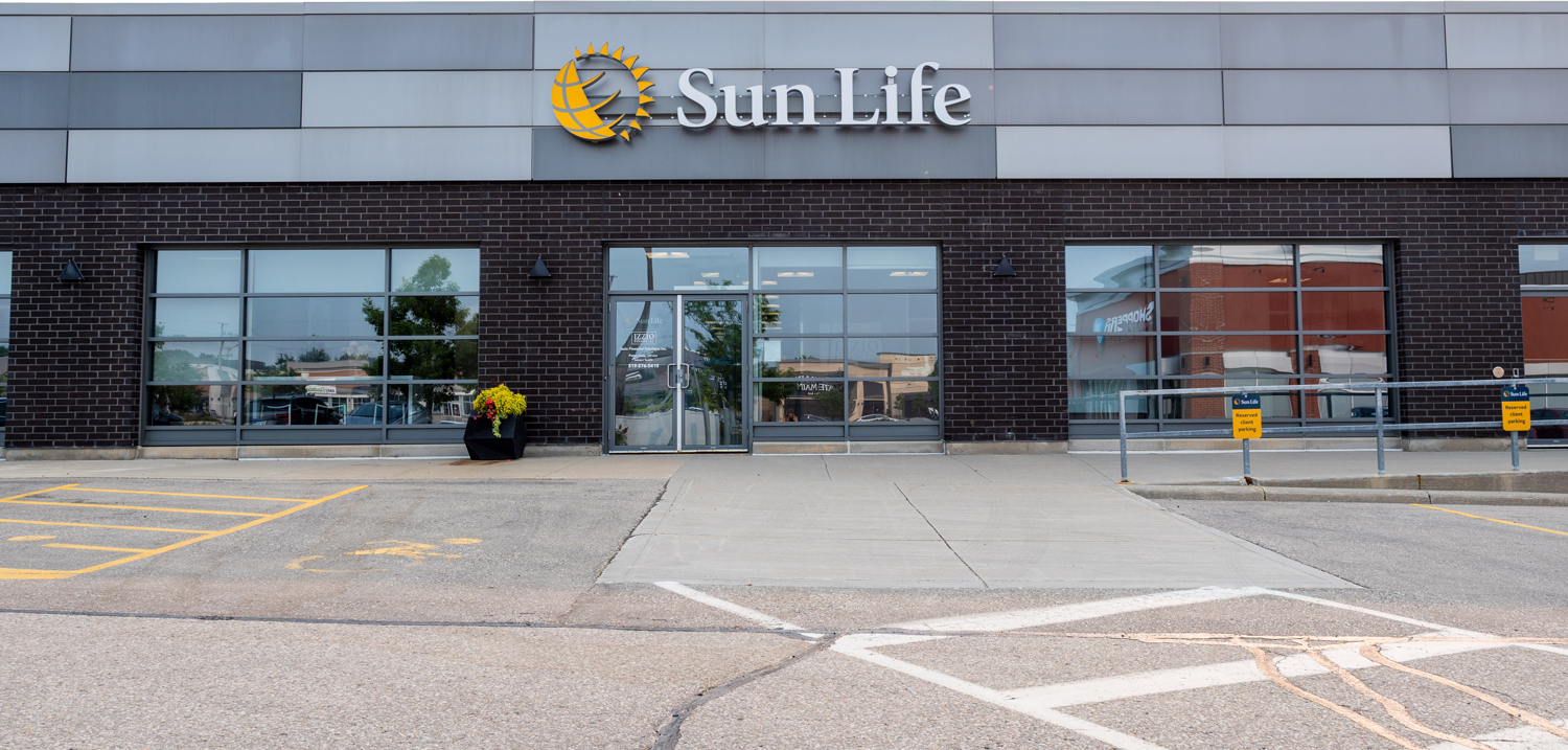 Exterior of the Sun Life office building with a dark brick facade, grey metal panels, and the Sun Life logo above glass doors and windows. A yellow 'Reserved client parking' sign hangs on a railing in front.