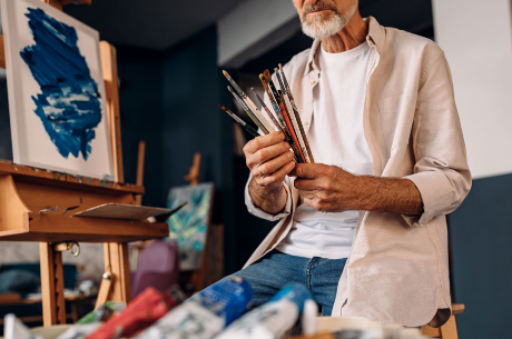 Older man in a casual shirt holding paintbrushes while sitting in an art studio.