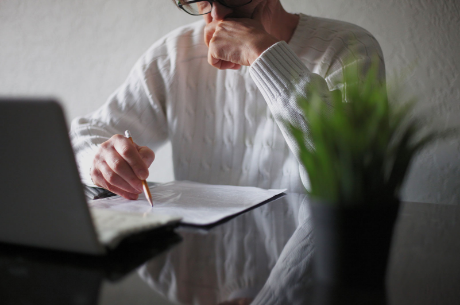 Person in a white sweater writing on paper with a pencil, with a laptop and a plant on the table.