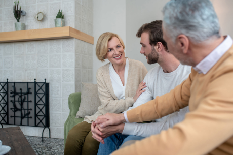 Older woman smiling and talking with a younger man and an older man, sitting on a couch in a cozy living room.