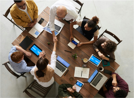  A group of people sitting around a table conducting a business meeting with laptops while two people shake hands.