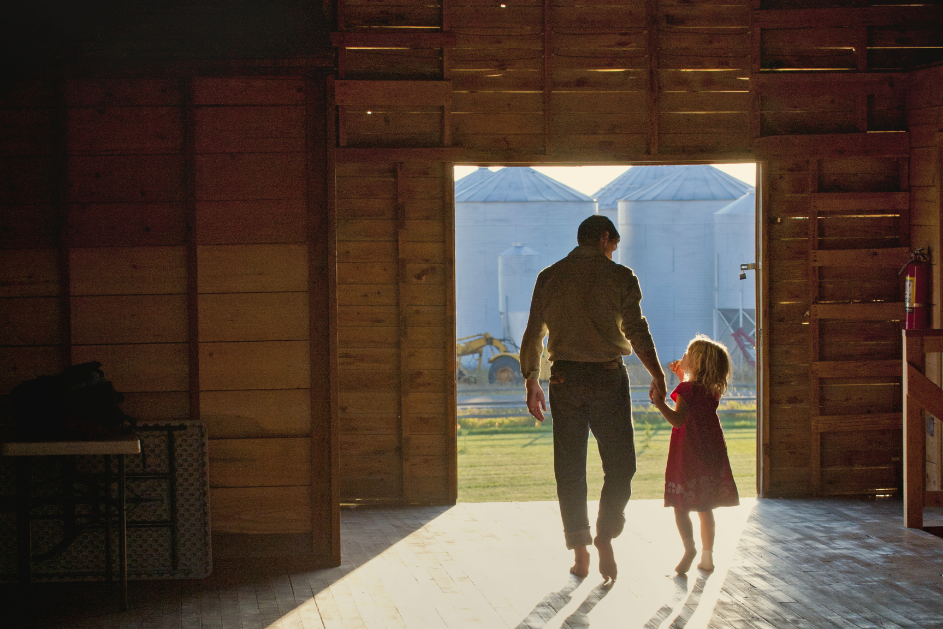 Man holds the hand of a girl as they walk out of a barn into a farmyard.